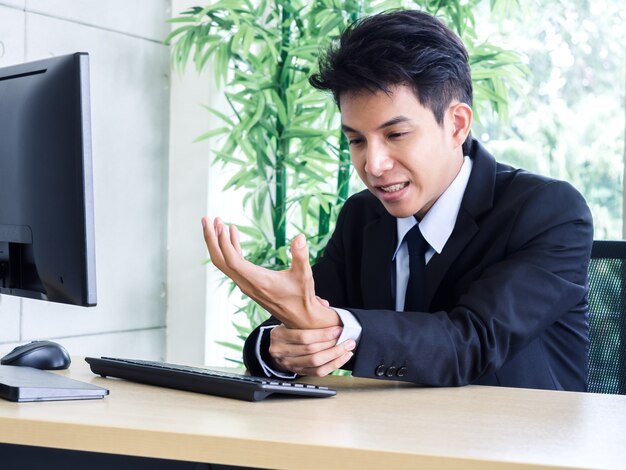 スーツを着た若いアジア人ビジネスマンがオフィスでノートパソコンを使用中に手の痛みを感じる