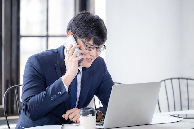 스마트폰과 노트북 컴퓨터 작업 및 화상 회의를 사용하는 젊은 아시아 사업가, 노트북을 사용하고 휴대 전화로 이야기하는 젊은 아시아 창의적인 남자.