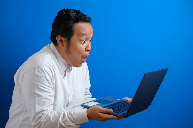 Young Asian businessman wearing casual white shirt looking at laptop, surprised expression. Close up body portrait