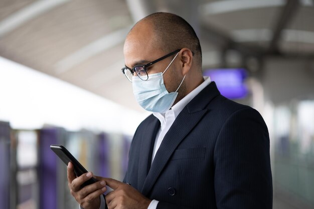 Young asian businessman wearing protection mask using smartphone standing at platform skytrain station. Businessman in urban city travel by sky train.