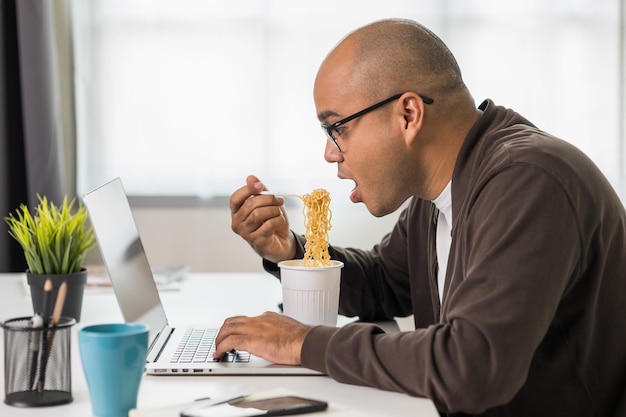 仕事中にインスタントラーメンを食べるラップトップインドのフリーランサーと一緒に働く若いアジア人実業家