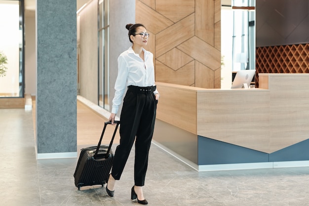 Молодая азиатская бизнес-леди в строгой одежде тянет чемодан в ожидании администратора в холле отеля