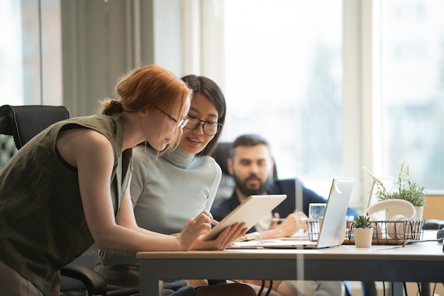 Young Asian businesswoman and her Caucasian colleague looking at display of touchpad while consulting about points of report