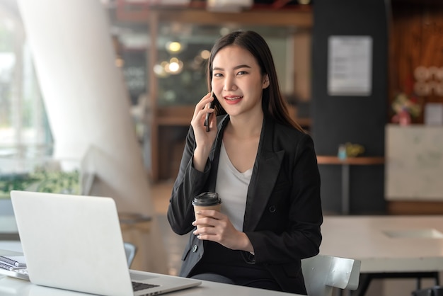 電話で話しているコーヒーカップを保持している若いアジアの実業家。オフィスでカメラを見てください。