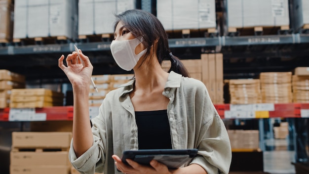 Young asian businesswoman manager wearing face mask warehouse using digital tablet checking inventory