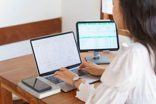 Young asian businesswoman reading marketing data of business on tablet and typing information on laptop while analysis strategy about investment and planning of new startup project to working in cafe