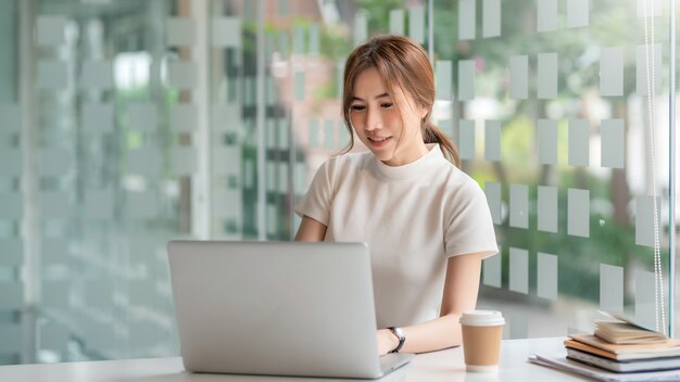 Young Asian businesswoman sitting at a modern office working on a laptop coffee at the table.