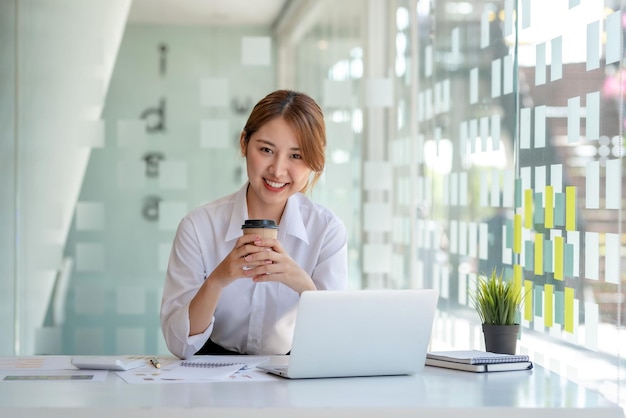 Молодая азиатская деловая женщина делает перерыв на кофе во время работы в офисе, глядя в камеру