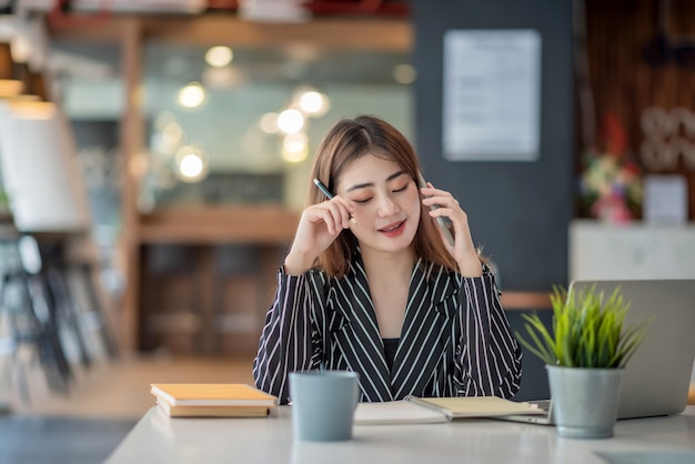 Young asian businesswoman talking on mobile phone working on laptop In modern office