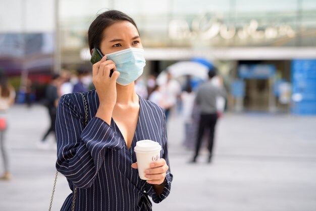 新しい通常の屋外として外出先でコーヒーを飲みながら電話で話しているマスクを持つ若いアジアの実業家