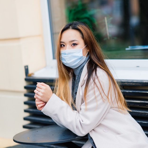 Giovane imprenditrice asiatica con maschera utilizzando il telefono e seduto a distanza al di fuori della caffetteria