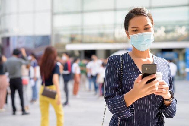 Young Asian businesswoman with mask using phone while having coffee on the go as the new normal during covid-19