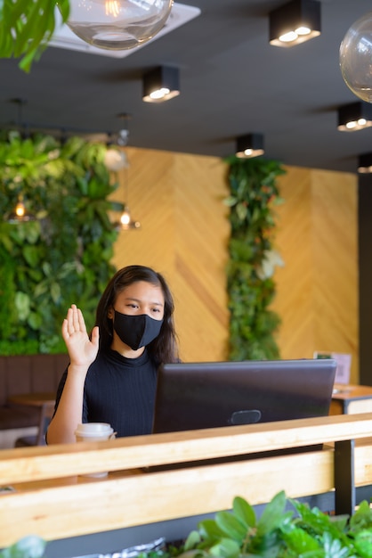 Giovane imprenditrice asiatica con videochiamata maschera e seduta con distanza all'interno della caffetteria