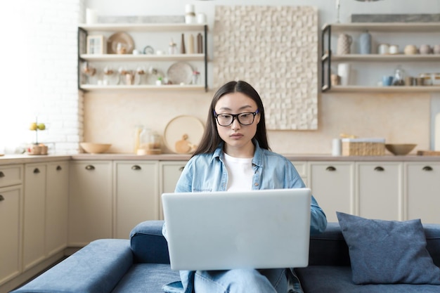 Young asian businesswoman working remotely from home sitting on sofa serious and focused using