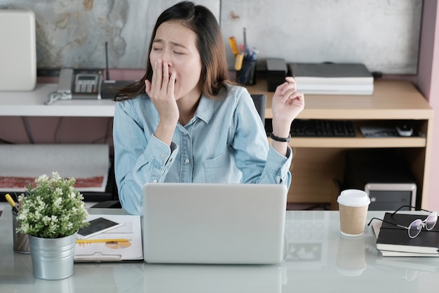 Молодой азиатский бизнесмен, зевая при работе с ноутбуком в офисе