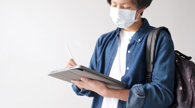 Giovane studente asiatico del campus che indossa una maschera di protezione mentre studia online con tablet digitale nel campus, prevenzione del coronavirus all'università, distanziamento sociale, apprendimento online