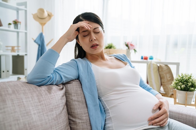 Young asian chinese pregnant woman with headache sitting on sofa couch in living room. Pregnancy symptoms expectation parenthood concept. lady at home holding belly baby feeling hurt at home.