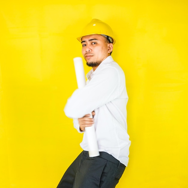 A young asian civil engineer in a hard hat is standing on a\
yellow background