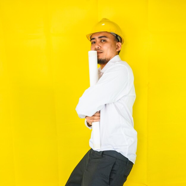 A young asian civil engineer in a hard hat is standing on a\
yellow background