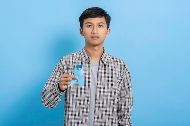 Young asian college student carrying books and backpack serious expression holding blue ribbon isolated over blue background