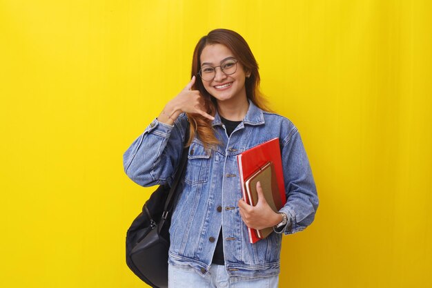 Young asian college student standing while holding books with calling hand gesture