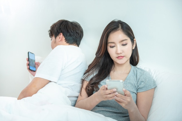 Giovani coppie asiatiche a letto facendo uso delle parti posteriori del telefono che si trovano l'un l'altro l'uomo e la donna dell'asia usano il proprio telefono cellulare astuto con segretezza, problema di relazione con la tecnologia.