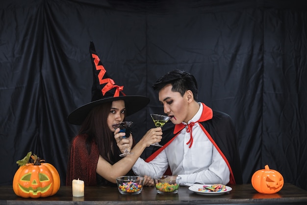 Giovane coppia asiatica in costume da strega e dracula con festeggiare la festa di halloween per un bicchiere tintinnio e bere nel festival di halloween. le coppie in costume celebrano il fondo del panno nero della festa di halloween.