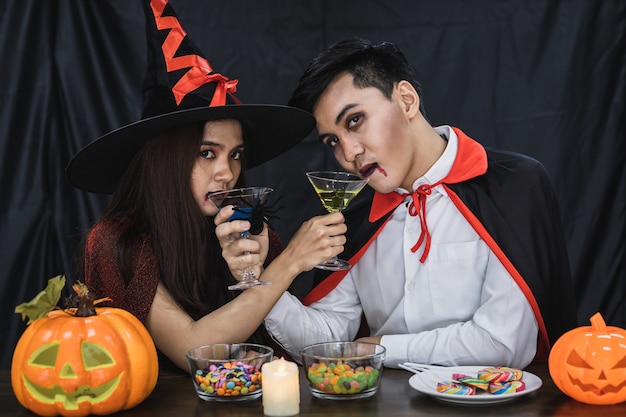 Young Asian couple in costume witch and dracula with celebrate Halloween party for clink glass and drink in Halloween festival. Couple in costume celebrate Halloween party black cloth background.