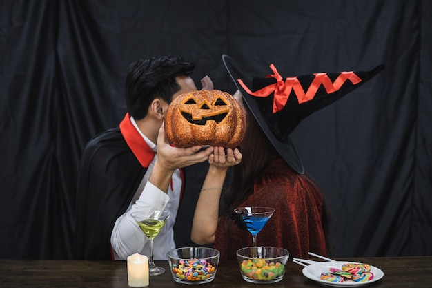 Foto giovane coppia asiatica in costume strega e dracula con festeggiare la festa di halloween e baciata con una zucca che copre il viso. le coppie in costume celebrano il fondo del panno nero della festa di halloween.