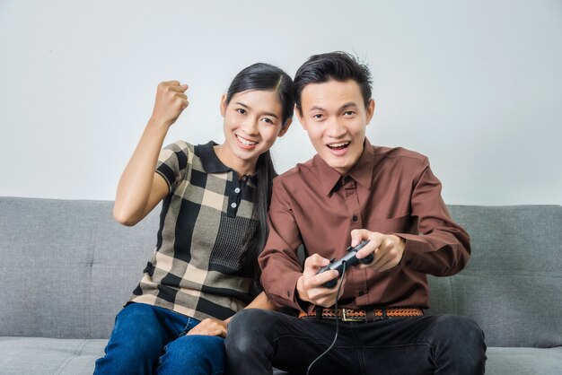 Young asian couple in love and playing video games holding joystick while sitting on sofa in living room.