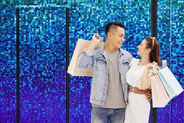 Молодая азиатская влюбленная пара стоит у сверкающей стены с хозяйственными сумками и смотрит друг на друга