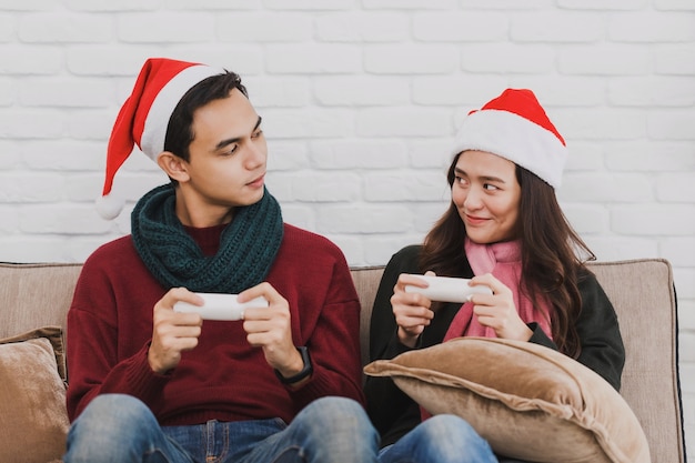 산타클로스 모자를 쓴 젊은 아시아 커플 연인. 집 배경에서 크리스마스 트리로 장식된 방에서 함께 비디오 게임을 하고 있습니다. 크리스마스 축하 컨셉입니다.