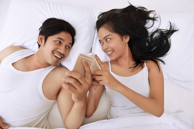 Young asian couple lying down in bed and using mobile phones