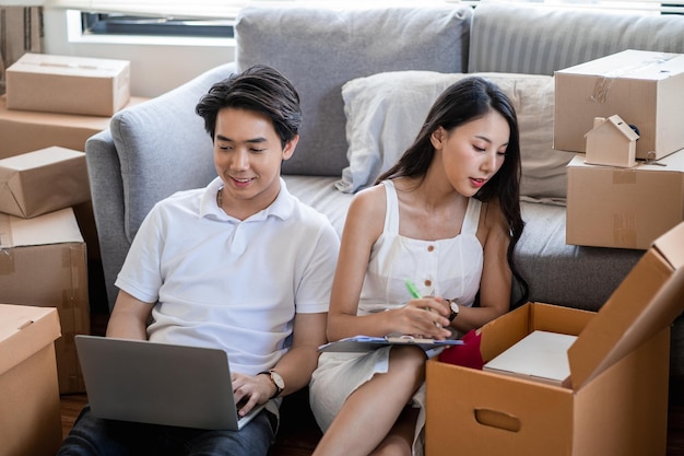 Young  asian couple moving to new home, sitting on the floor very happy and cheerful for new apartment around cardboard boxes and holding cardboard boxes while moving home