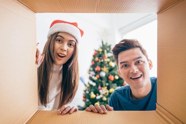 Young Asian couple opening a Christmas present together with a smile views from inside of the box with a decorated Christmas tree in the background Boxing day during the Christmas holiday