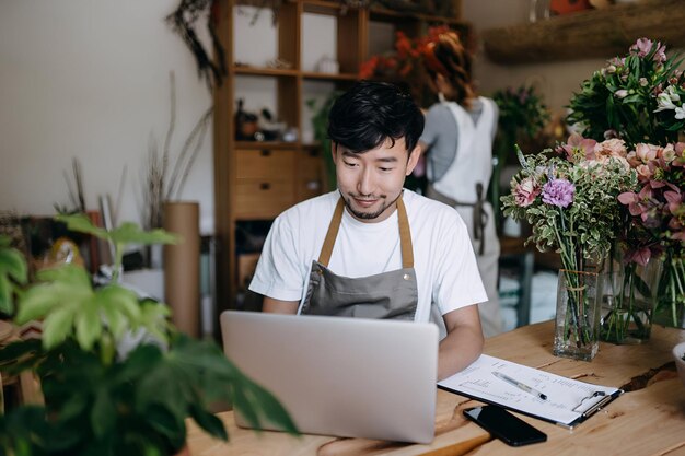 写真 花や植物の隣のカウンター越しにラップトップで作業する中小企業の花屋のアジア人の若いカップルのオーナーがオンラインで商品を販売する顧客の注文を受けて在庫を確認する