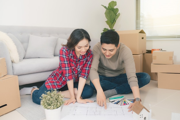 Young asian couple sitting on the floor and looking at the blueprint of new home.