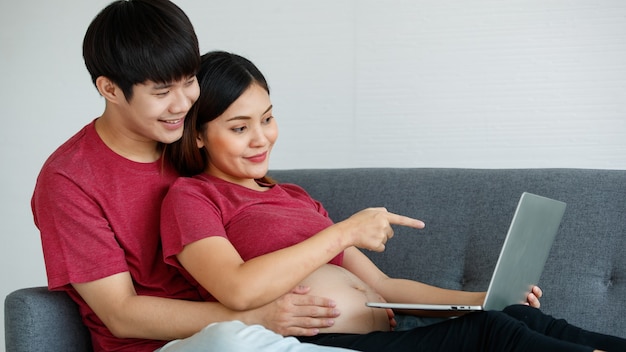 Young asian couple sitting relaxedly together on a couch. a\
pregnant woman point at a laptop screen while a man smile and puts\
his hands around her. family concept.