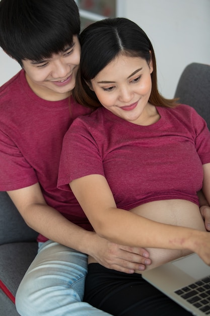 Young asian couple sitting relaxedly together on a couch. a\
pregnant woman point at a laptop screen while a man smile and puts\
his hands around her. family concept.