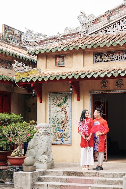 Young Asian couple in traditional clothes with peach branches and paper fan leaving Buddhist temple