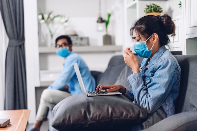 Giovani coppie asiatiche che utilizzano il funzionamento del computer portatile e la videoconferenza che incontrano la chat online nella quarantena per il coronavirus che indossa la maschera protettiva con l'allontanamento sociale a casa concetto del lavoro da casa