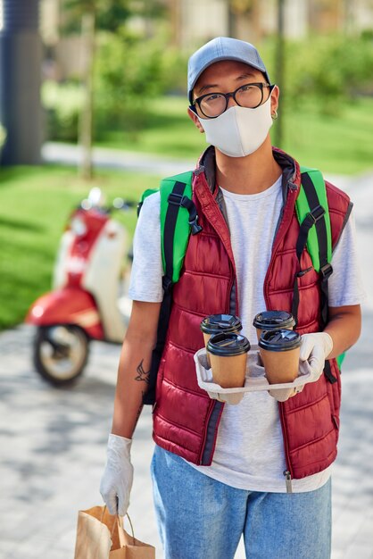 Young asian courier wearing face protective mask and gloves holding four coffee cups and paper
