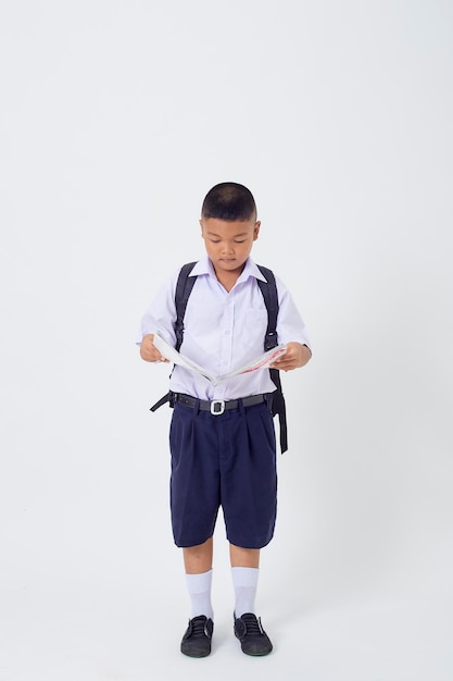 Foto un giovane ragazzo carino asiatico in piedi in un'uniforme scolastica thailandese con una borsa zaino e un libro su uno striscione bianco sullo sfondo