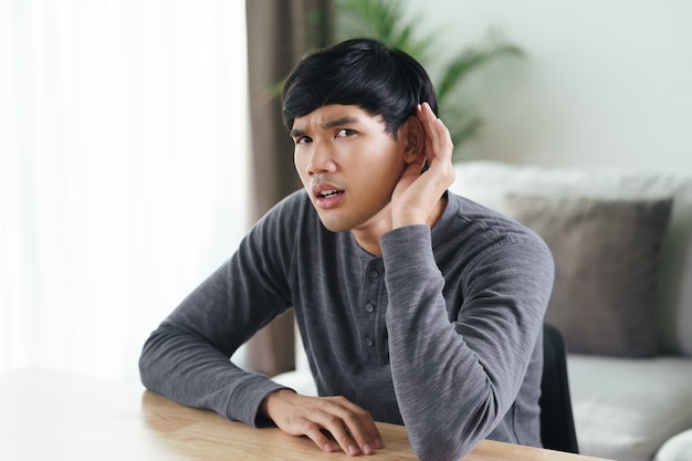 聴覚に問題のある若いアジアの聴覚障害者の男性は、耳に手をかざし、注意深く耳を傾け、難聴です。