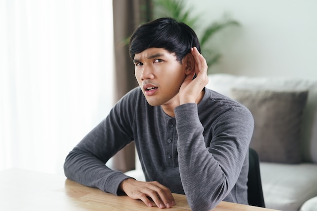 Young Asian deaf disabled man having hearing problems holds his hand over the ear, listens carefully, hard of hearing.