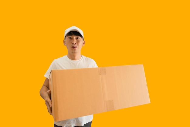 Photo young asian delivery man wearing cap and white blank tshirt holding parcel post box isolated on yellow background express delivery service concept