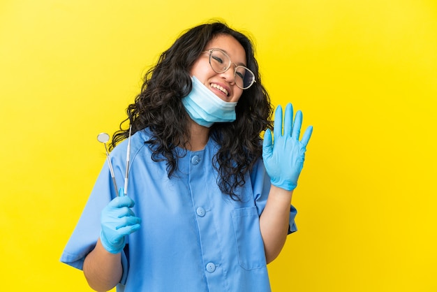 Giovane dentista asiatico che tiene gli strumenti su sfondo isolato salutando con la mano con espressione felice