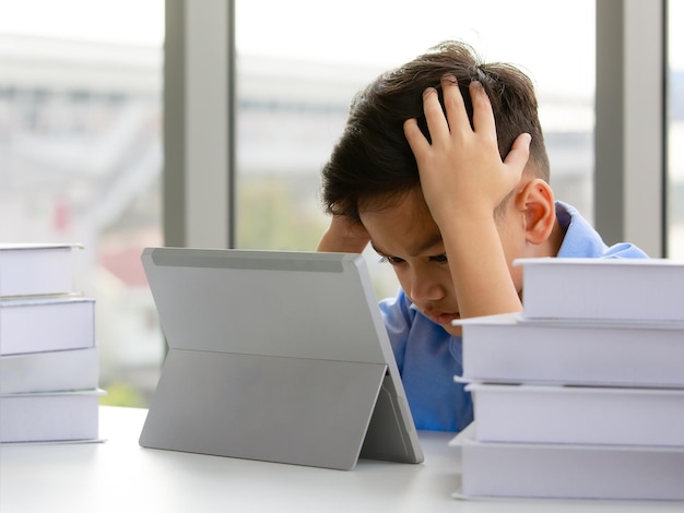 Young Asian elementary school boy sitting next to stack of books and using tablet computer with both hands holding his head with gestures and face showing stress