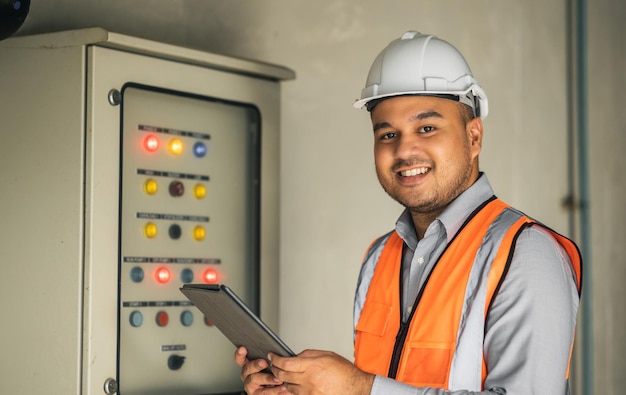 Giovane ingegnere asiatico che lavora al controllo e alla manutenzione dell'armadio elettrico per la sicurezza operaio che utilizza un tablet per controllare l'impianto elettrico dell'edificio