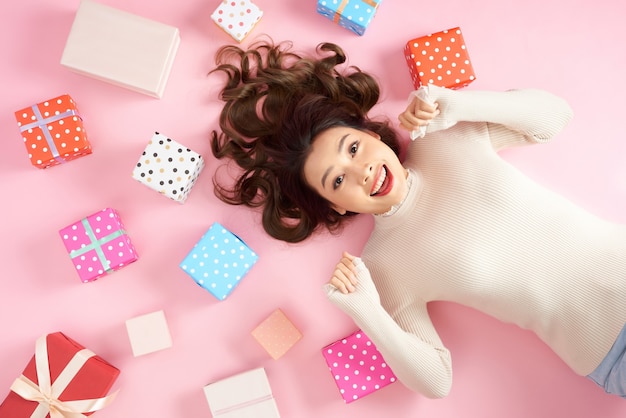 Young Asian excited woman lying on pink floor with many colorful gift boxes.